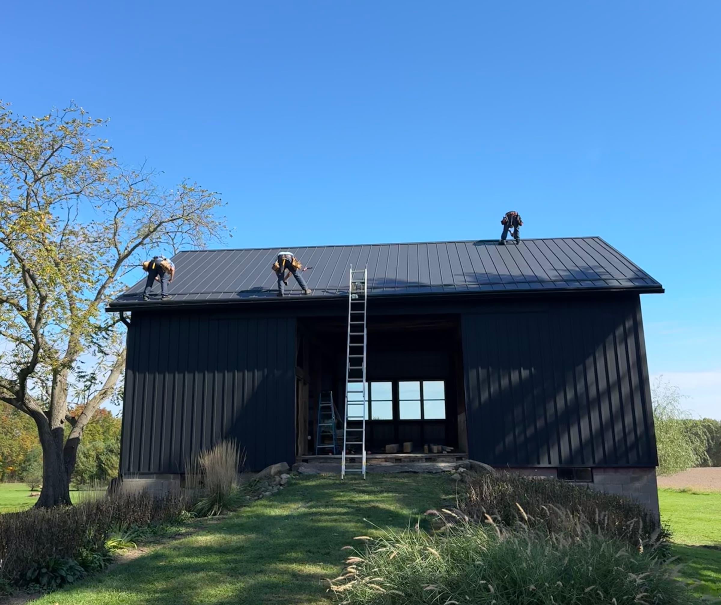 Crew installing standing seam metal roofing on a black board-and-batten barn